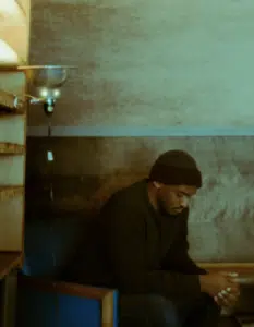 A person wearing a beanie and dark sweater sits in a chair, looking down with hands clasped. Shelves and a lamp are visible beside them.
